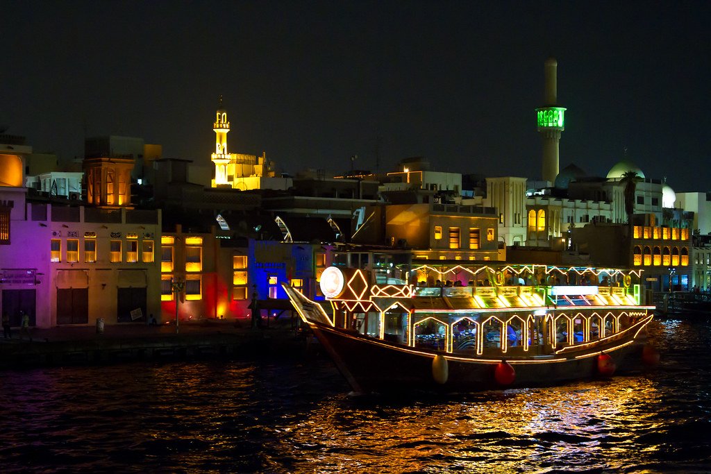 Illuminated traditional Dhow boat cruising on Dubai Creek at night with historic buildings and minarets lit up in the background.