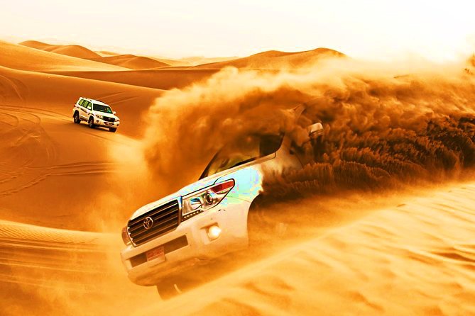White SUV dune bashing in Dubai desert safari, kicking up sand with another vehicle in the background.