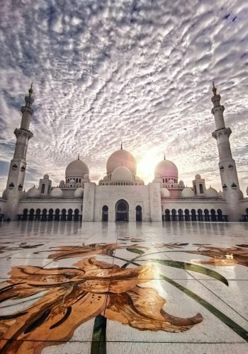 A low-angle, wide-angle photo of the Sheikh Zayed Grand Mosque in Abu Dhabi, UAE, featuring white marble domes and minarets against a cloudy sky with the sun in the background. The foreground shows an intricate floral mosaic on the courtyard floor.